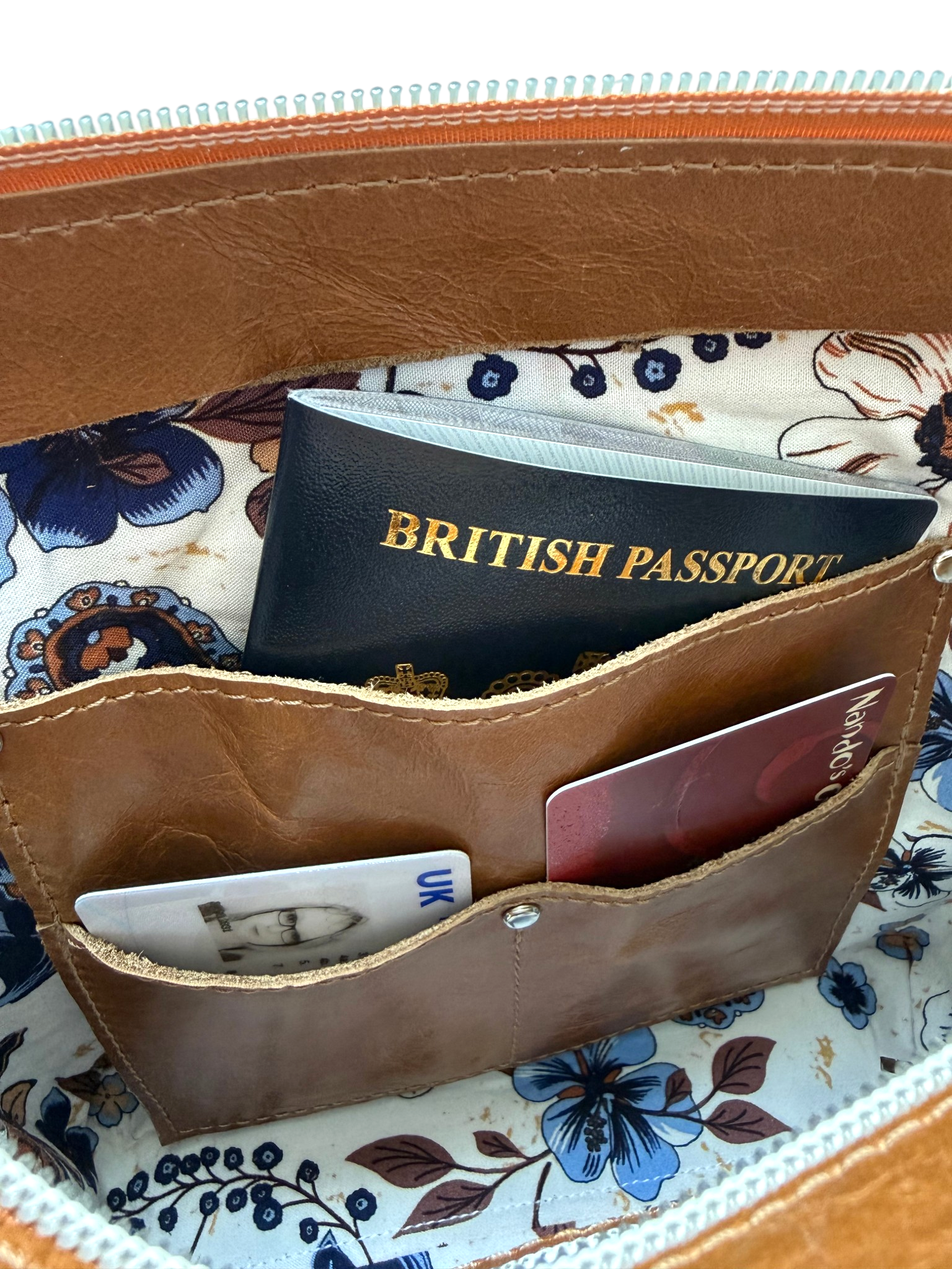 View of the integrated slip pocket inside a tan brown leather bag, showing a British passport and cards against a floral patterned lining