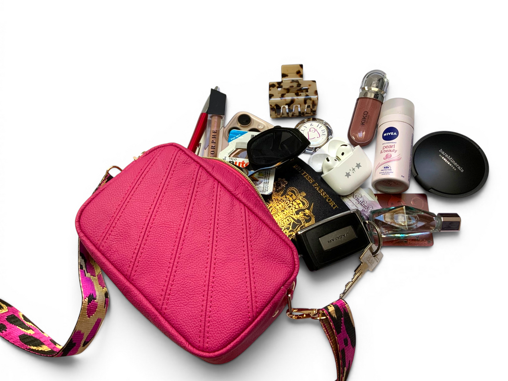View of a fuchsia pink crossbody bag with various beauty and travel products spilling out on a white background