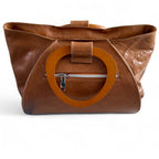 View of the back of a tan leather handbag with wooden handles, and zipper pocket, sitting on a white background.