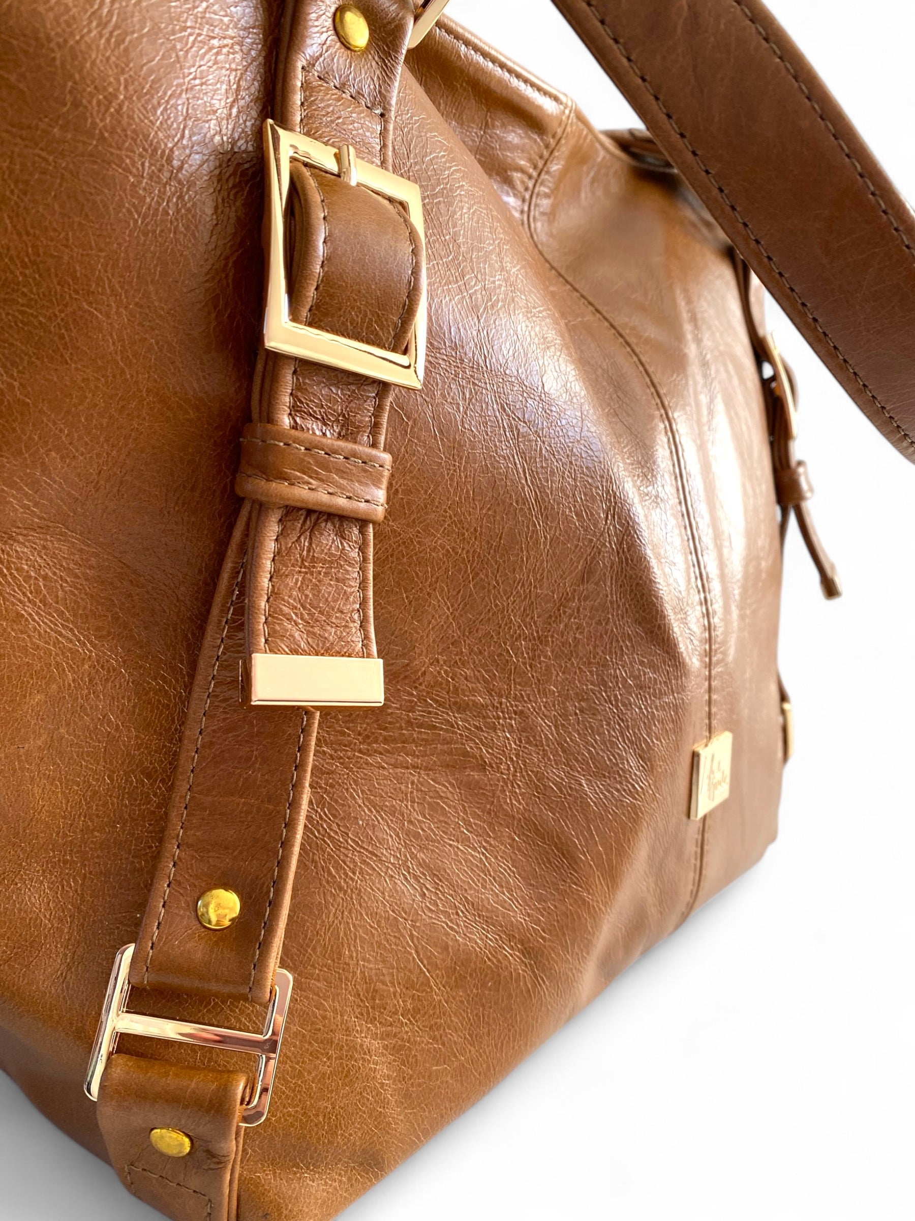 Closeup image of the details of a tan brown leather bag. It shows the gold hardware of a decorative strap
