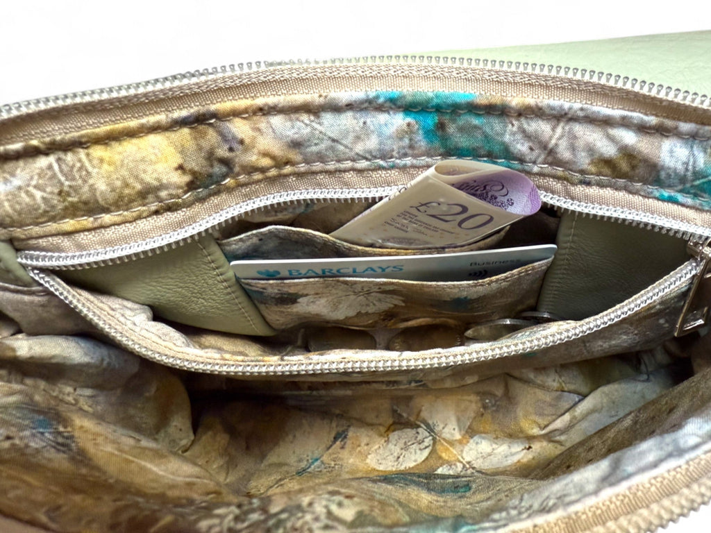 Image shows a pale green Olivia bag. The crossbody bag is open and is showing the inside of the interior zippered pocket, which serves as an integrated purse. There is a £20 note in the back section a bank card in the front card pocket and coins in the bottom of the pocket. 