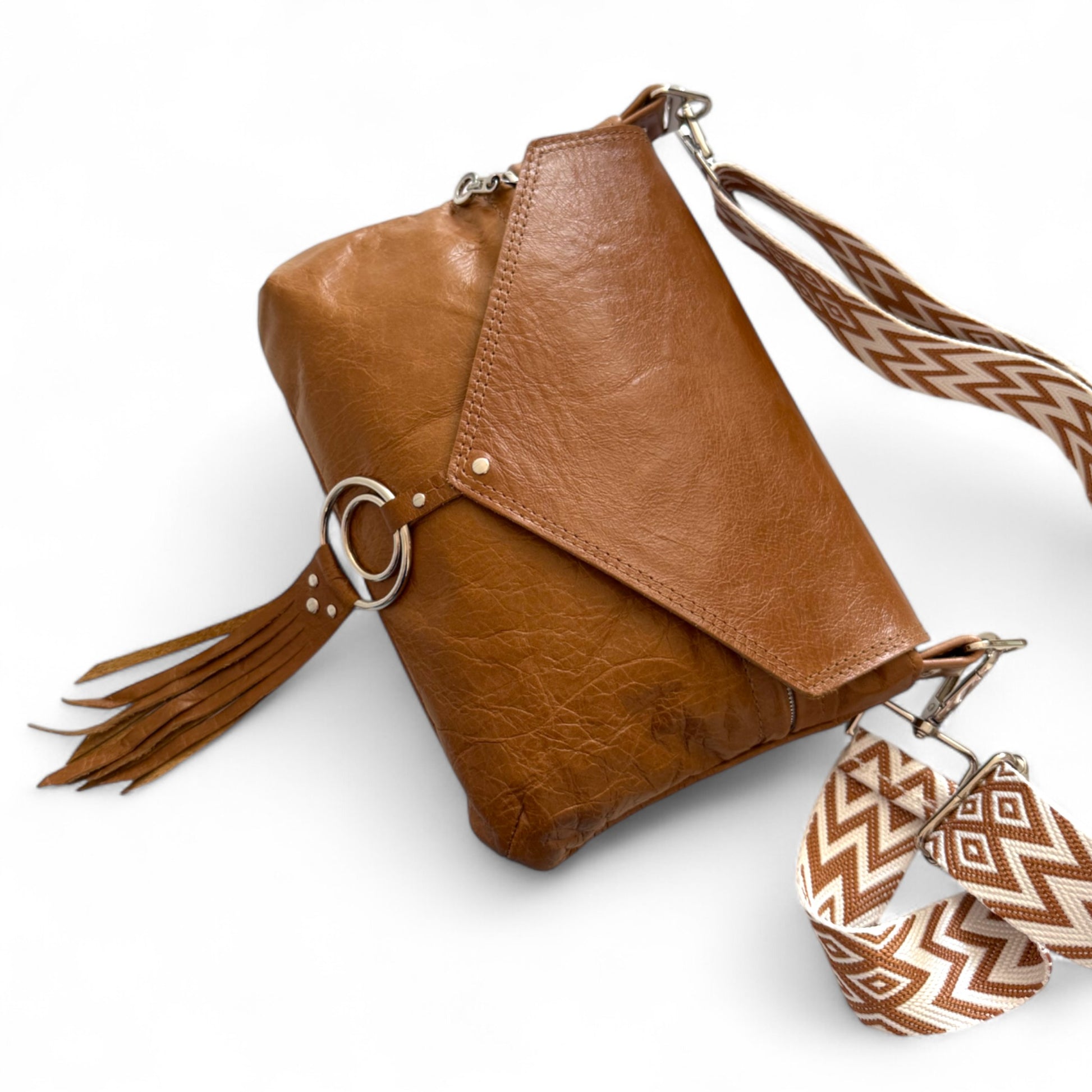 Tan Olivia crossbody bag, lying on a white background. The front of the flap has a double ring and leather fringe. The bag has a matching, chevron patterned crossbody strap attached. 