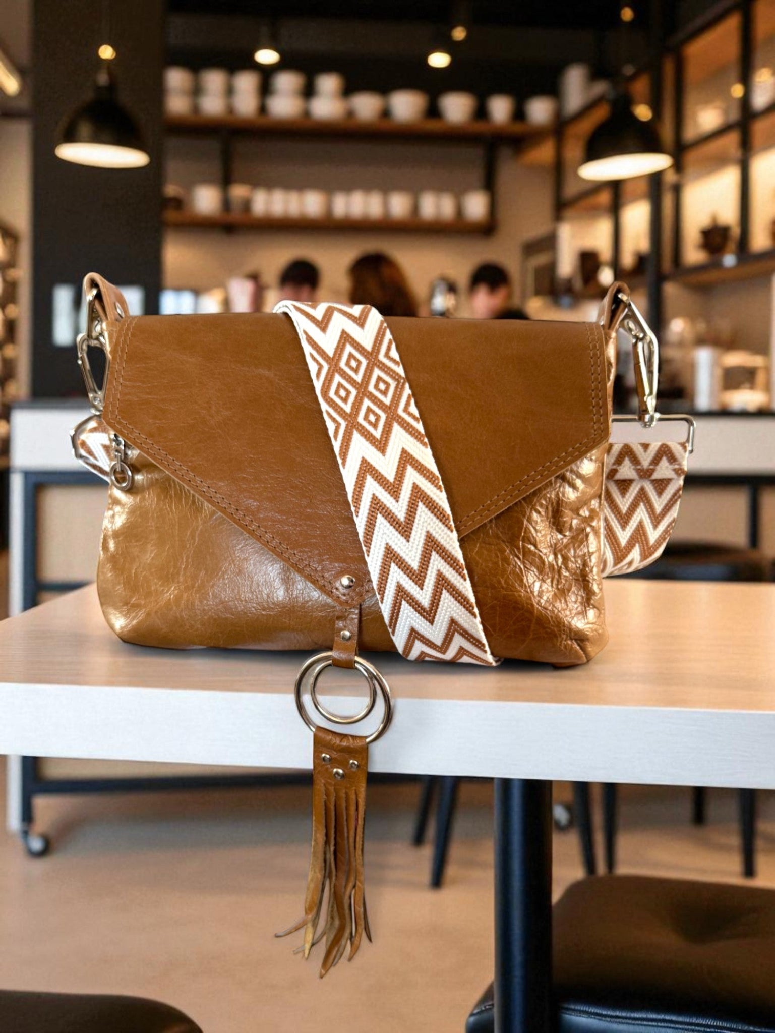 Tan brown handbag with a decorative strap on a table in a cafe setting. The Olivia crossbody bag is hand crafted using leather that has been rescued from heading to landfill. This bag is an exclusive, one-off bag by Lilly & Jude