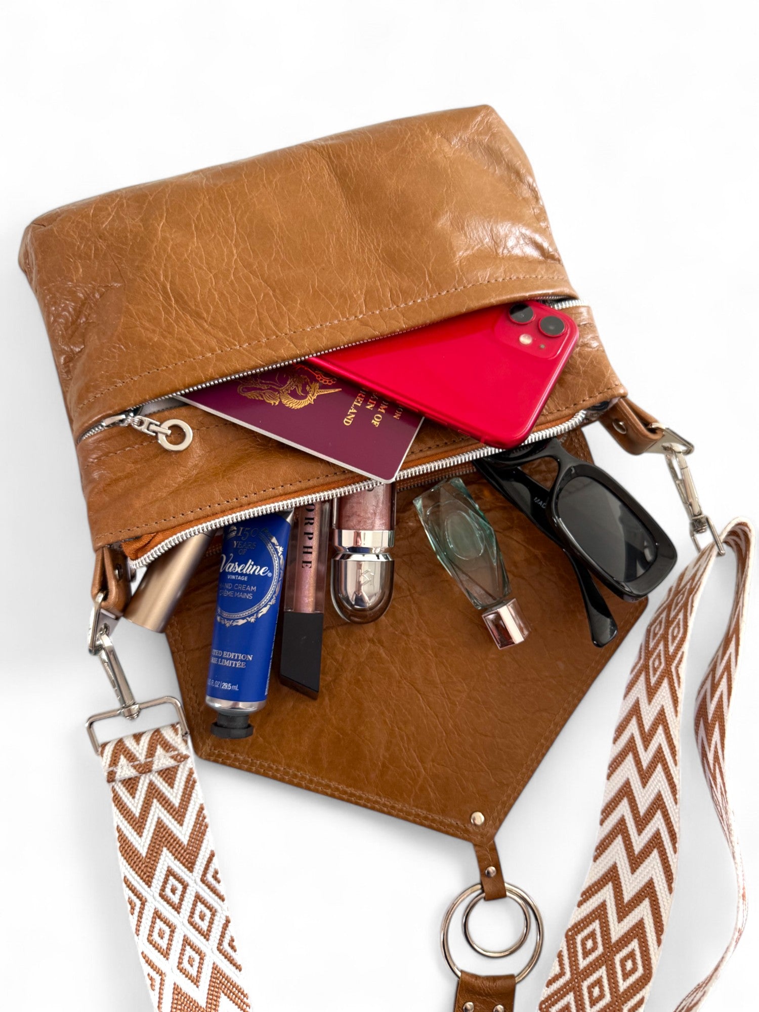 tan brown leather bag, lying down on a white background, with the contents, such as cosmetics, perfume and sunglasses spilling out. The front pocket contains a red mobile phone and a passport