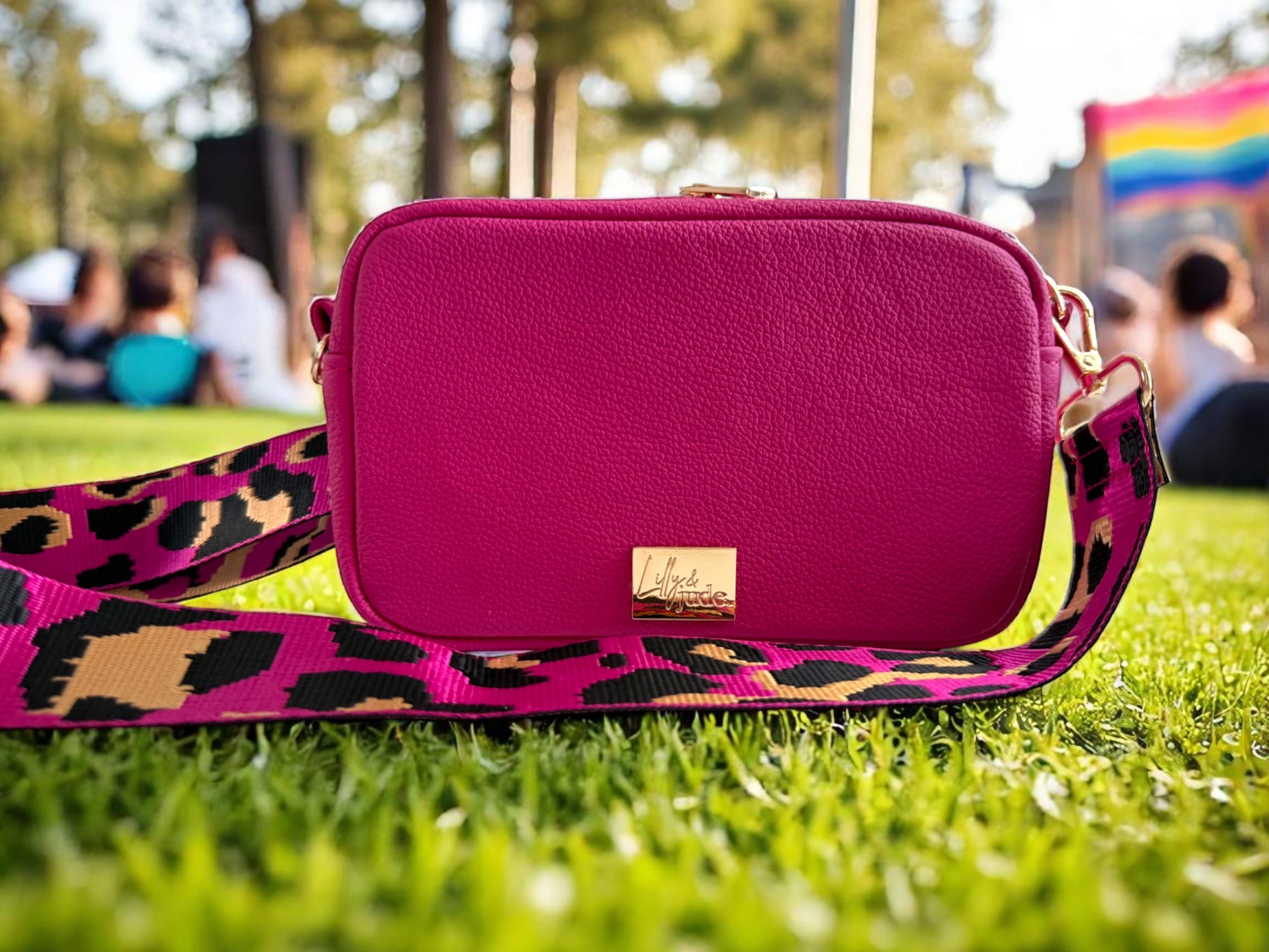Tiny Harriet hot pink compact crossbody bag by Lilly & Jude sitting on grass at a festival with a blurred pride flag in the background.